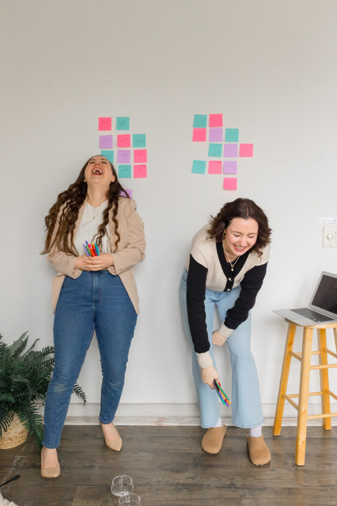 Two women standing in front of a wall filled with post-it notes. Both are holding colorful pens and laughing.