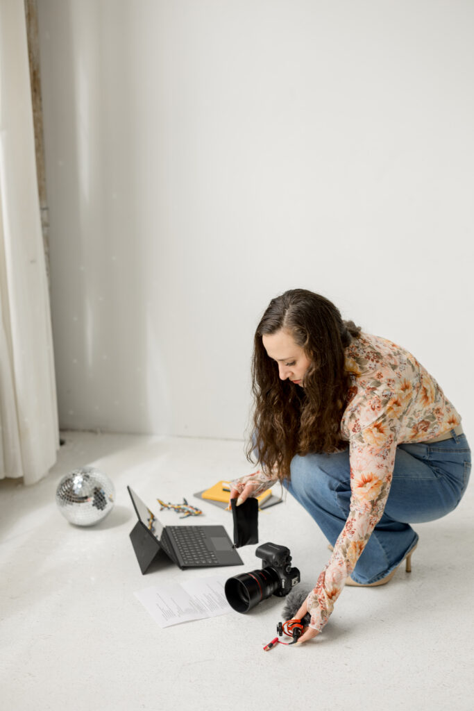 woman organizing microphones on the floor next to a computer, a shot list, a camera, a stack of notebooks and planners, and a couple of decorative items.