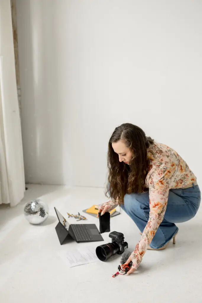 woman organizing microphones on the floor next to a computer, a shot list, a camera, a stack of notebooks and planners, and a couple of decorative items.